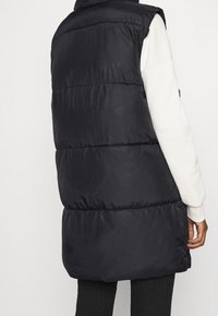 Svart vadderad väst med hög krage, med horisontella quiltade sektioner, som når till mitten av låret, bärs över en ljusfärgad långärmad skjorta.