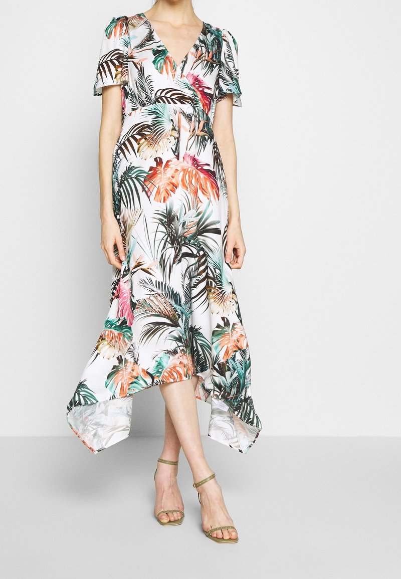 Femme portant une robe blanche à col en V avec imprimé coloré de feuilles tropicales et ourlet asymétrique, associée à des talons à bride cheville vert olive.