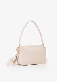 Pequeño bolso rectangular beige con un solo mango superior, cremallera dorada y dos etiquetas de cuero adjuntas sobre un fondo blanco.