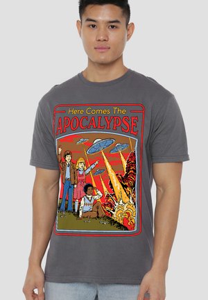 Young man wearing gray t-shirt showing cartoon of children observing UFOs firing beams causing explosions, with text "Here Comes The Apocalypse."