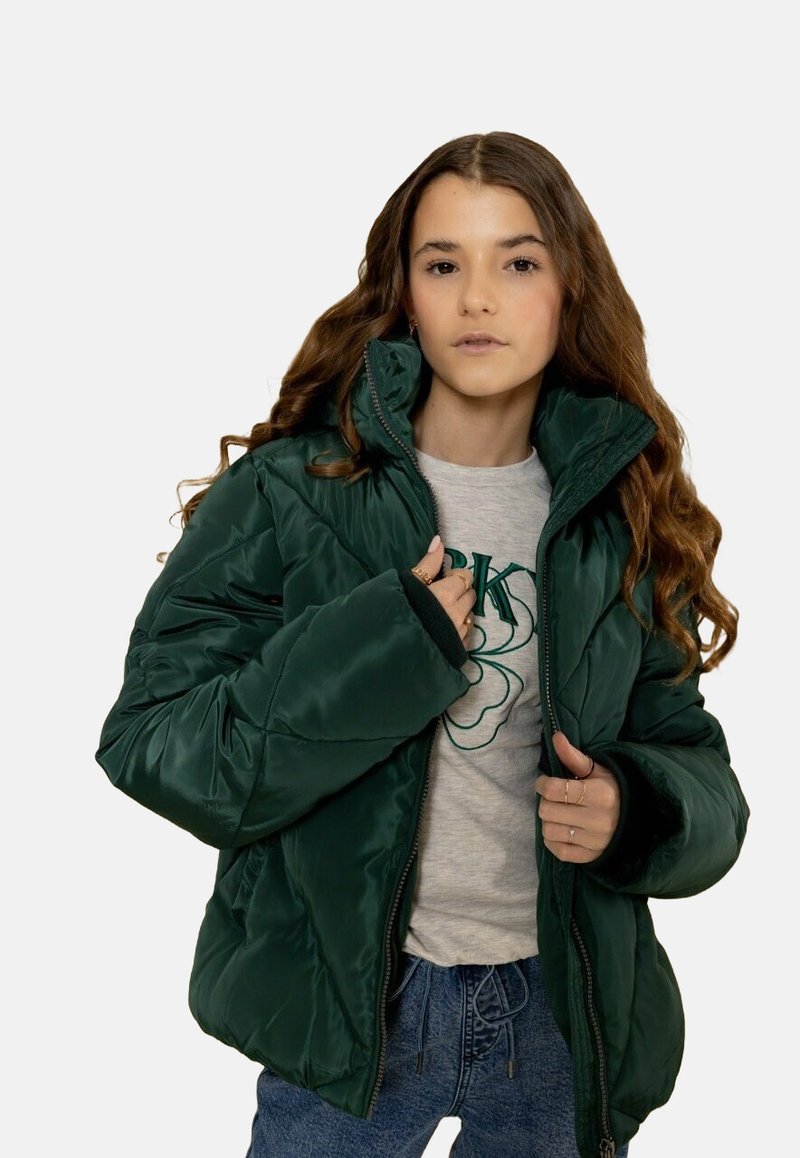 Donkergroen pufferjack met een hoge kraag, ritssluiting en een gestructureerd oppervlak. Gecombineerd met een grijs shirt en een blauwe spijkerbroek.