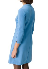 Femme portant une robe bleu clair mi-longue à manches longues, avec une couture dans le dos et de petites fentes aux poignets sur fond blanc.