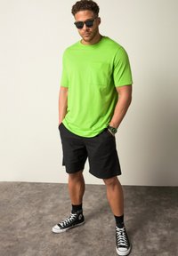 Neonově zelené tričko s krátkým rukávem a kapsou na hrudi, spárované s černými šortkami a černými vysokými teniskami. Matný povrch, volný střih.