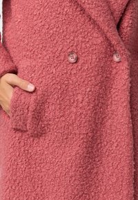 Manteau rose texturé avec une surface douce et duveteuse, doté de deux boutons ronds et d'une poche latérale. Design classique à double boutonnage.