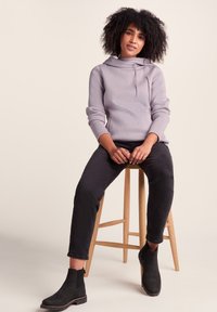 Light purple sweatshirt with a cowl neck and drawstrings, paired with black slim-fit jeans and black ankle boots. Wooden stool background.