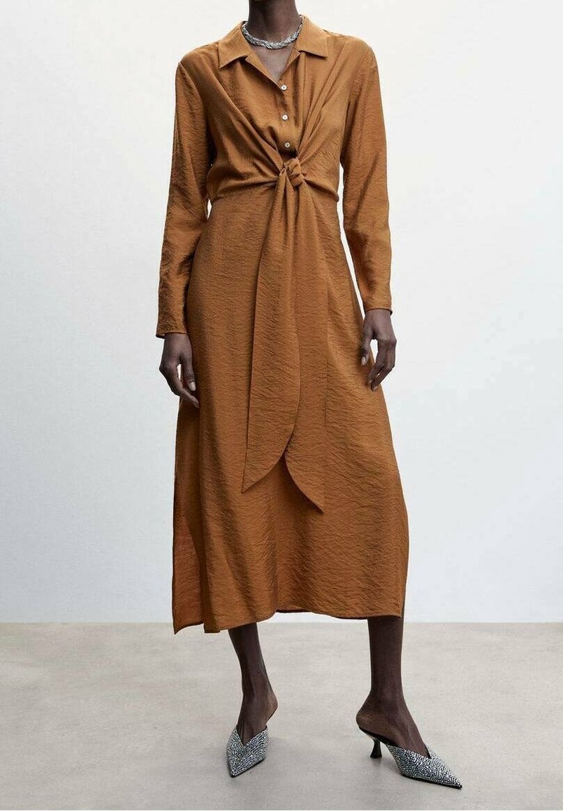 Femme portant une robe midi à manches longues couleur rouille avec un détail de nœud à l'avant, associée à des mules à talons hauts pointues à motif.