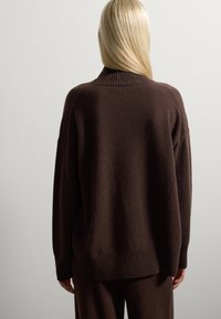 Persoon met lang blond haar, gekleed in een losse donkerbruine trui en bijpassende broek, gezien vanaf de achterkant tegen een effen witte achtergrond.