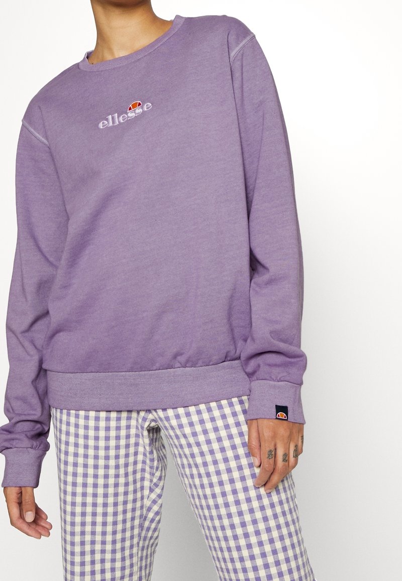 Personne portant un sweat-shirt ellesse violet et un pantalon à carreaux violet et blanc, montrant une partie de sa main droite tatouée.