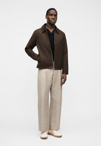 Bărbat purtând o jachetă maro cu fermoar, tricou negru, pantaloni largi bej și adidași albi, stând pe un fundal alb simplu.