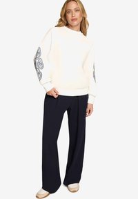 Femme portant un sweat-shirt blanc avec des motifs cœur paisley bleus sur les manches, associé à un pantalon noir à jambes larges et des baskets blanches.