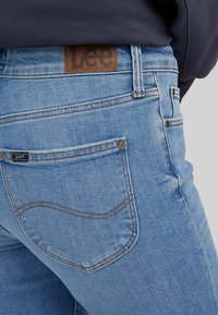 Vaaleansiniset farkut, joissa on ruskea nahkaleima vyötäröllä. Ominaisuuksiin kuuluu tavalliset taskut ja kontrastivärinen tikkaus.