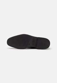 Black rubber shoe sole with a textured surface, featuring ridges for grip and a logo accent. Shape is flat with a rounded toe.