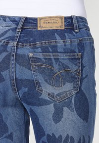 Jeans en denim d'un ton bleu moyen avec un motif floral. Caractéristiques d'un design de poche arrière distinctif et d'une étiquette en cuir marquée au-dessus de la taille.