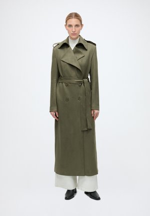 Donna in piedi di fronte a noi, che indossa un trench verde oliva con cintura sopra pantaloni bianchi a gamba larga e scarpe nere con punta su sfondo bianco.