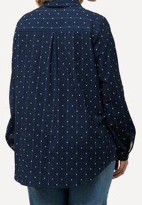 Navy blue shirt with white polka dots, long sleeves, button cuffs, rear pleat, and curved hem. Cotton material, relaxed fit.