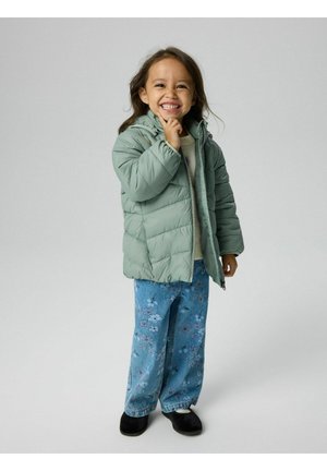 Jeune fille souriante portant une doudoune verte, un jean à fleurs et des chaussures noires, debout devant un fond clair uni.