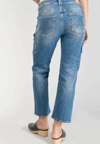 Vista trasera de una persona que lleva jeans ajustados azules con bolsillos traseros y un bolsillo de cargo lateral, combinados con zuecos negros de cuña sin talón.