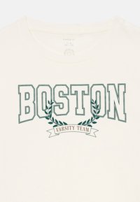 Krémové tričko s výrazným zeleným nápisem "BOSTON" nad "VARSITY TEAM" na banneru, doplněné vavřínovými listy. Měkký bavlněný materiál.