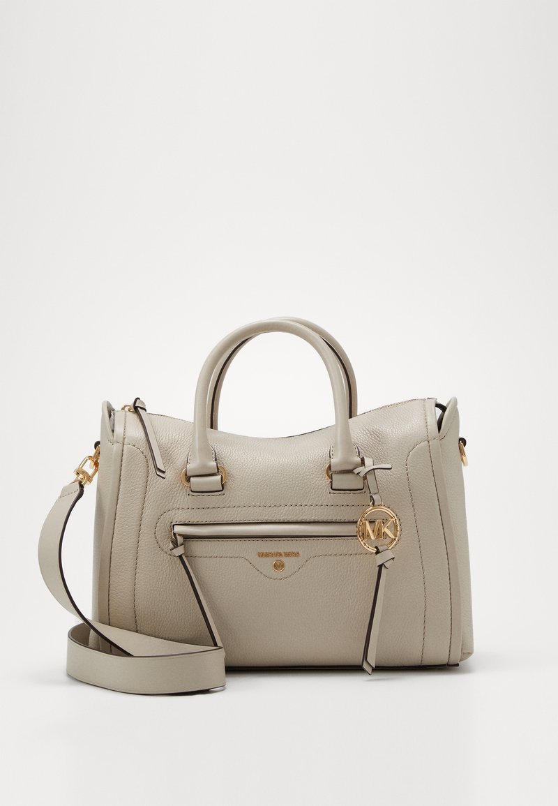 Jasnobeżowa skórzana torebka z dwoma uchwytami, regulowanym paskiem na ramię, przednią kieszenią na zamek i złotym zawieszonym logo "MK" na jednolitym tle.