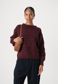 Maglione lavorato a maglia color bordeaux con motivi tessuti, scollo rotondo e vestibilità ampia; abbinato a una borsa a tracolla marrone e pantaloni neri.