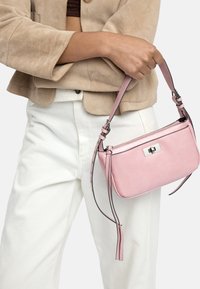 Beżowa kurtka i białe spodnie noszone przez osobę trzymającą małą różową torebkę z srebrnym zapięciem i regulowanym paskiem.