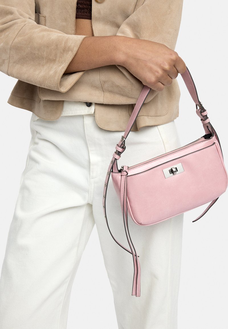 Beżowa kurtka i białe spodnie noszone przez osobę trzymającą małą różową torebkę z srebrnym zapięciem i regulowanym paskiem.