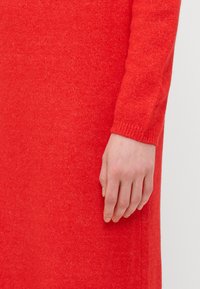 Abito rosso a maglia con una texture liscia, manica aderente e polsini a coste. L'attenzione è posta sul colore vivace e sui dettagli del tessuto.