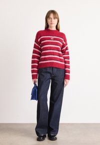 Rood en roze gestreepte sweater met een geribbelde halslijn, gecombineerd met donkere blauwe wijde jeans. Accessoires zijn een kleine blauwe handtas.