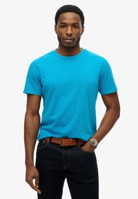 Kortærmet turkise T-shirt lavet af bomuld, båret med mørke jeans og et brunt bælte. Har rund hals og afslappet pasform.