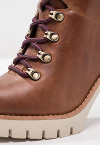 Bottine en cuir marron avec œillets en métal, lacets bleu marine et rouge, et semelle épaisse beige. Présente un talon légèrement surélevé et une surface texturée.