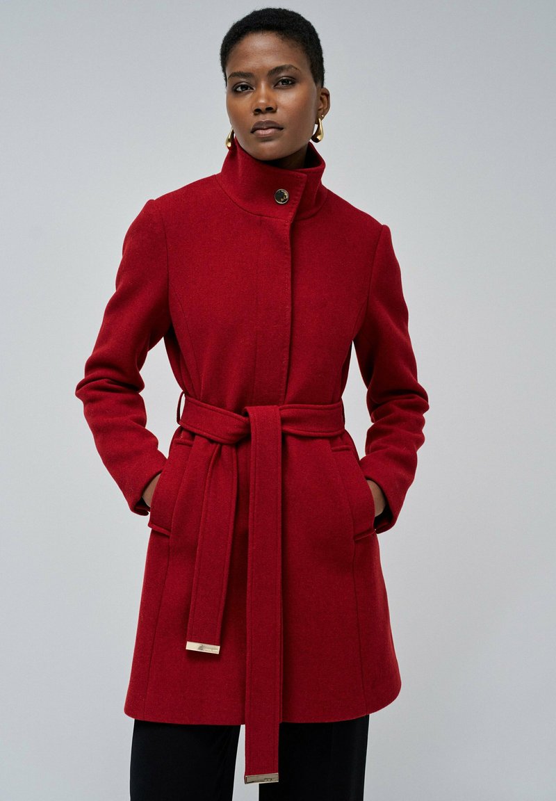 Crvena kaput od vune s visokim ovratnikom, strukiranim pojasom i dva prednja džepa. Ima glatku teksturu i metalne akcentne detalje na remenu.