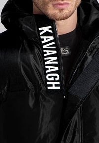 Černá prošívaná bunda s lesklou texturou, která má kontrastní bílé logo "KAVANAGH" v oblasti zipu a pevný zip.