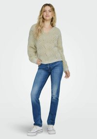 Hellgrüner Pullover mit einem strukturierten, wellenförmigen Muster, kombiniert mit blauen Jeans und weißen Sneakers. Der Pullover hat einen V-Ausschnitt und einen lockeren Sitz.
