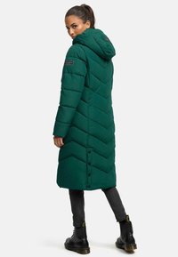 Cappotto imbottito verde lungo con cappuccio, presenta un motivo a chevron, bottoni a pressione e due tasche laterali. Indossato sopra pantaloni neri attillati e stivali.
