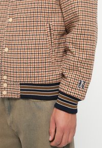 Brun og beige houndstooth-mønstret jakke med rund krage, knappefeste, ribbestrikkede mansjetter og hem som har marineblå og brune striper.