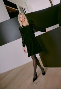 Sort halvgennemsigtig kjole med struktureret diamantmønster, lange ærmer og flæset kant, kombineret med gennemsigtige sorte strømpebukser og spidse sorte hæle.