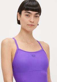 Μωβ μαγιό με λεπτές τιράντες και λογότυπο στο στήθος. Το ύφασμα φαίνεται λείο με ελαφριά γυαλάδα, διαθέτοντας εφαρμοστό σχέδιο.