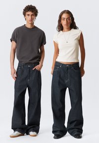 Dva modely nosí oversized, tmavě modré džíny s ohrnutými manžetami. Mužský model má na sobě tmavě šedé tričko s krátkým rukávem, zatímco ženský model má fitted krémový top.