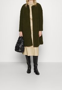 Cappotto verde oliva con texture e grandi bottoni, indossato sopra un vestito camicia beige. Abbinato a stivali neri al ginocchio e una borsa nera intrecciata.