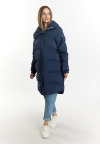 Geacă puffer de culoare albastră închisă cu guler înalt, cu secțiuni cusute, închidere cu fermoar și manșete ribbate. Asortată cu blugi deschiși și adidași albi.