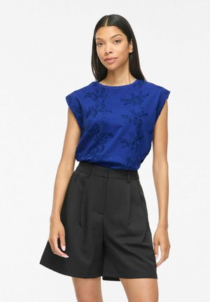 Femme portant un top bleu brodé sans manches et un short noir plissé taille haute, debout devant un fond blanc uni.