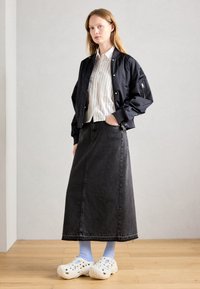 Femme portant une veste bomber noire, une chemise rayée blanche, une longue jupe en denim noire, des chaussettes bleues claires et des chaussures blanches à enfiler, se tenant à l'intérieur.