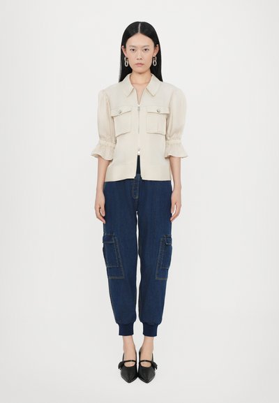 Beige zip-front blouse with short puff sleeves and two chest pockets, paired with dark blue cargo pants featuring side pockets and ribbed cuffs.