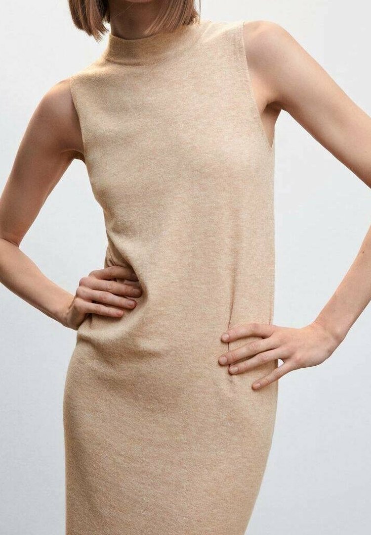 Femme portant une robe beige en maille sans manches, mains sur les hanches, sur un fond clair uni.