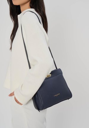 Frau trägt weißen Pullover und Hose und hält eine dunkelblaue Lancaster Paris Leder-Umhängetasche mit goldener Schnalle und Logo.