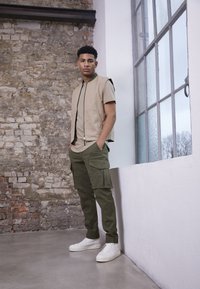 Colete bege sem mangas sobre uma camisa castanha, combinado com calças cargo verdes e ténis brancos. Apresenta fecho frontal e bolsos laterais.