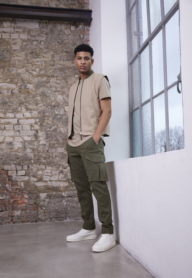 Colete bege sem mangas sobre uma camisa castanha, combinado com calças cargo verdes e ténis brancos. Apresenta fecho frontal e bolsos laterais.