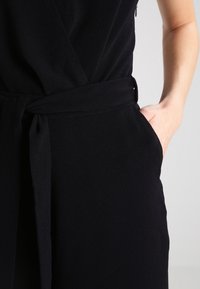 Svart byxdress med en invecklad design, tyget visar en fin struktur. Knytt bälte i midjan och fickor synliga på sidorna.