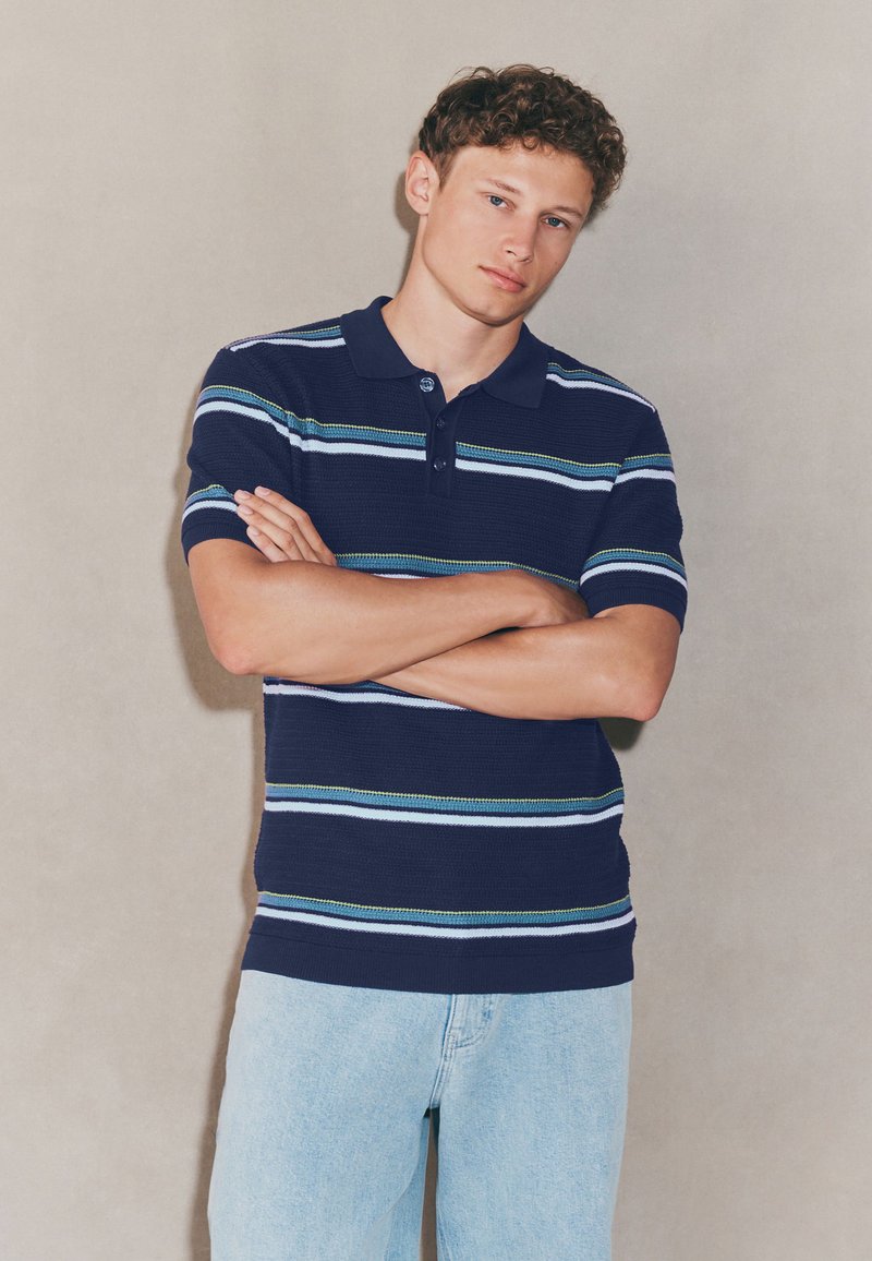 Tricou polo cu mânecă scurtă, cu dungi orizontale albastre și verzi, asortat cu blugi albastru deschis.