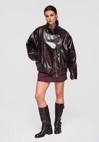 Kobieta w oversize'owej, błyszczącej ciemnobrązowej skórzanej kurtce, krótkiej bordo sukience i ciemnobrązowych kozakach za kolano stojąca na jednolitym tle.